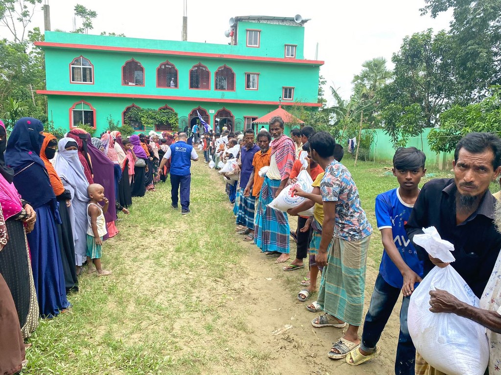 बांग्लादेश में बाढ़ राहत अभियान के दौरान आईओएम कार्यकर्ताओं से खाद्य सहायता प्राप्त करने के लिए पुरुषों, महिलाओं और बच्चों का एक समूह बाहर एक पंक्ति में खड़ा है।