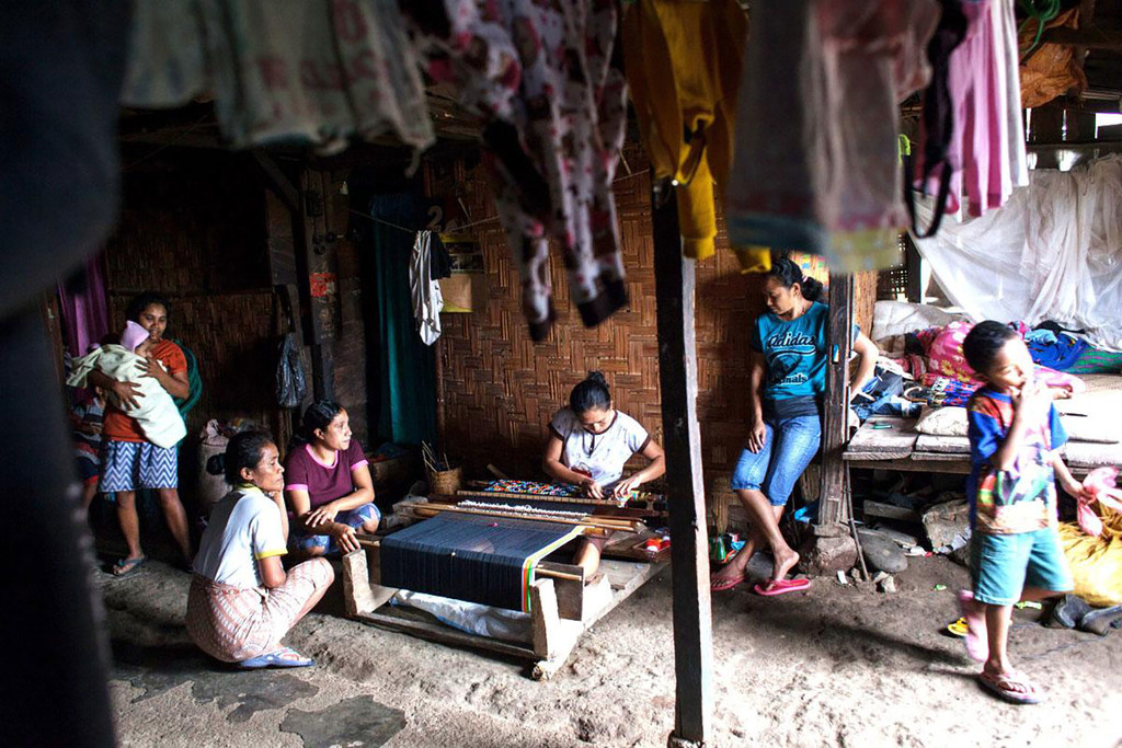 Un grupo de mujeres y un niño se encuentran en una vivienda rural, con una mujer tejiendo en un telar.