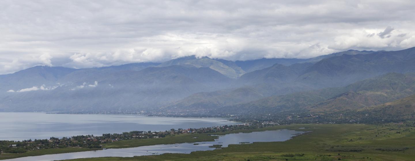 Vue panoramique du lac Kivu avec la ville d'Uvira sur la rive, dominée par des montagnes brumeuses sous un ciel nuageux.
