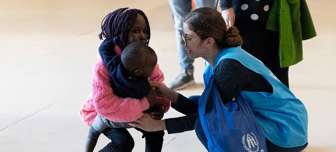 À l'aéroport de Lisbonne, un membre du personnel du HCR , vêtu d'un gilet bleu, s'accroupit pour saluer un jeune enfant et un tout-petit.