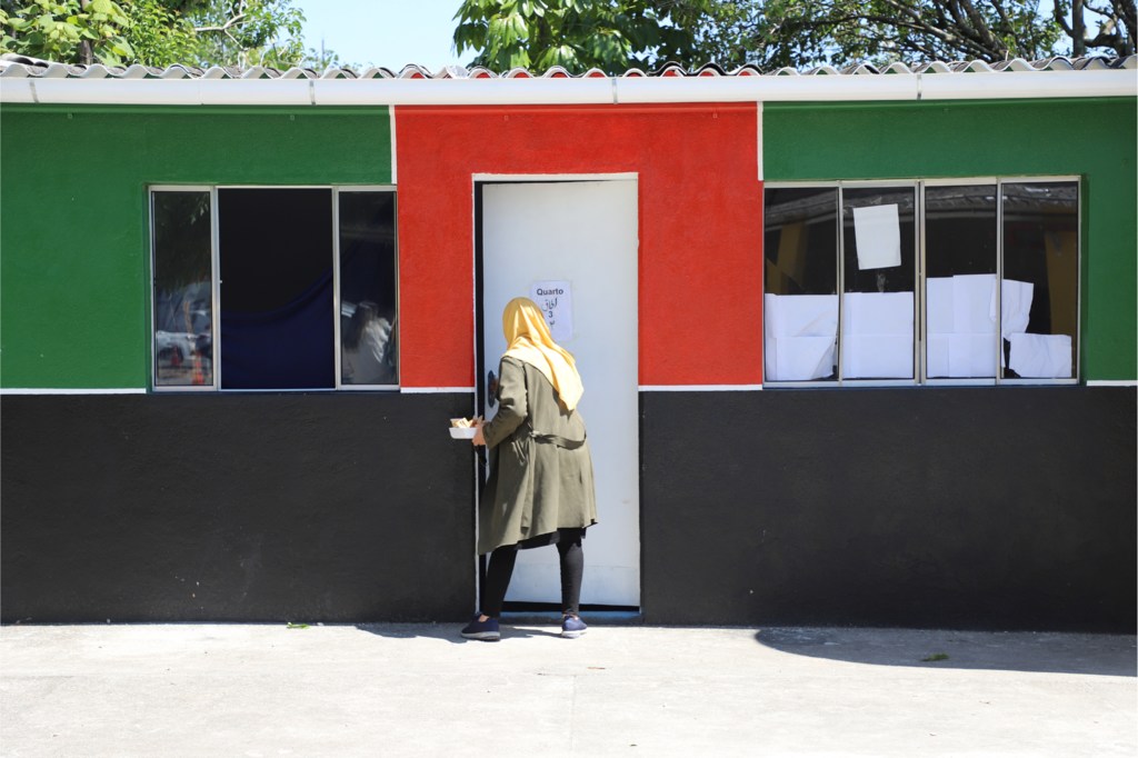 Uma mulher usando hijab está parada na entrada de um prédio colorido, segurando um pequeno recipiente.