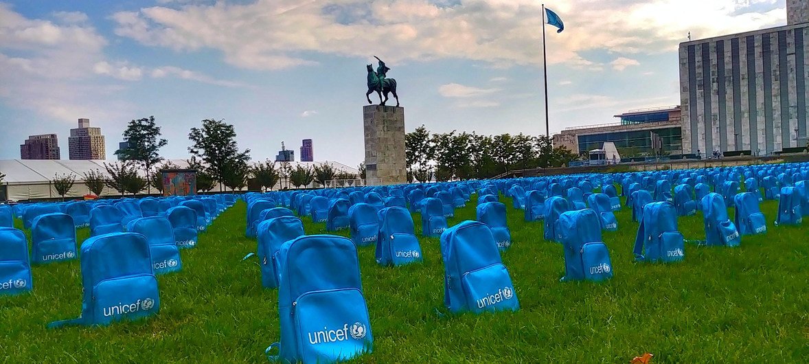 Fileiras de mochilas azuis da Unicef estão expostas em um gramado em frente a uma grande estátua e um prédio moderno.