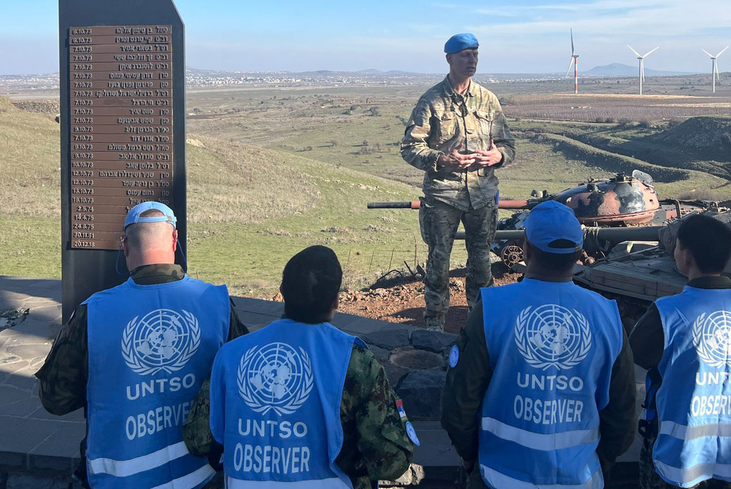 المراقبون العسكريون في السترات الزرقاء يقفون بالقرب من لوحة تذكاري مع التواريخ، ويستمعون إلى ضابط في الزي العسكري يتحدث. يمكن رؤية دبابة ومحركات الرياح في الخلفية.