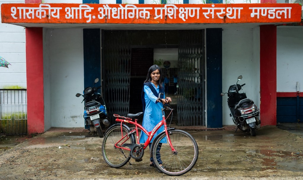 भारत में एक सरकारी व्यावसायिक प्रशिक्षण केंद्र की इमारत के सामने भारतीय लड़की याचना अपनी लाल साइकिल के साथ खड़ी है।
