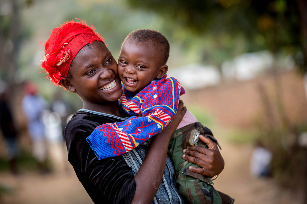 Mama Mwafrika aliyevaa kitambaa chekundu kichwani amemshikilia mtoto mchanga mwenye tabasamu akiwa amevaa sweta yenye rangi nyingi katika mazingira ya nje huko Blantyre, Malawi.
