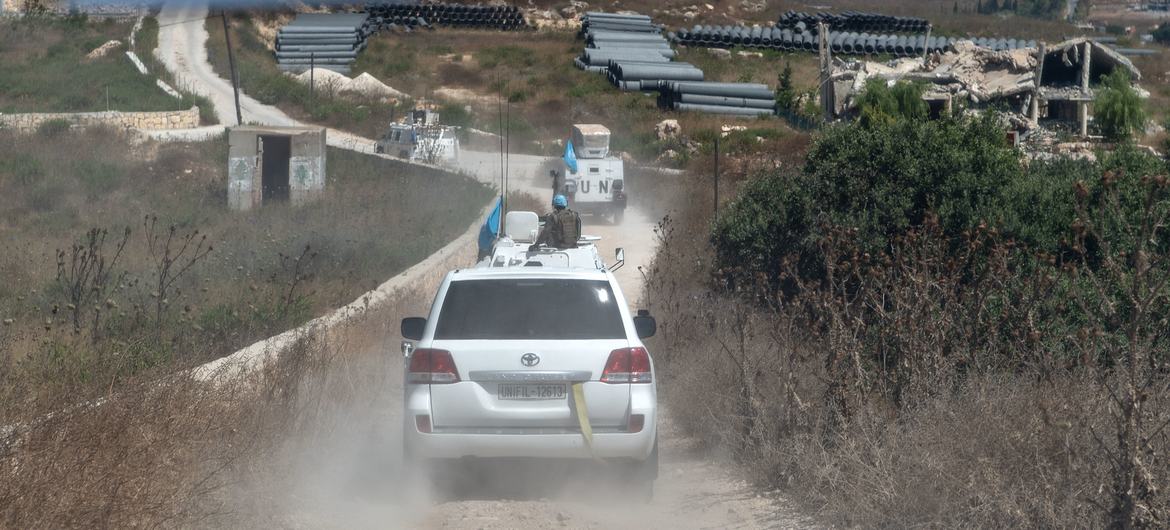 تسير مركبات UNIFIL ، بما في ذلك سيارة تويوتا لاند كروزر بيضاء وشاحنة تحمل أعلام الأمم المتحدة الزرقاء، على طول طريق ترابي في منطقة ريفية قاحلة مع وجود مواد بناء ومبانٍ متضررة في الخلفية.