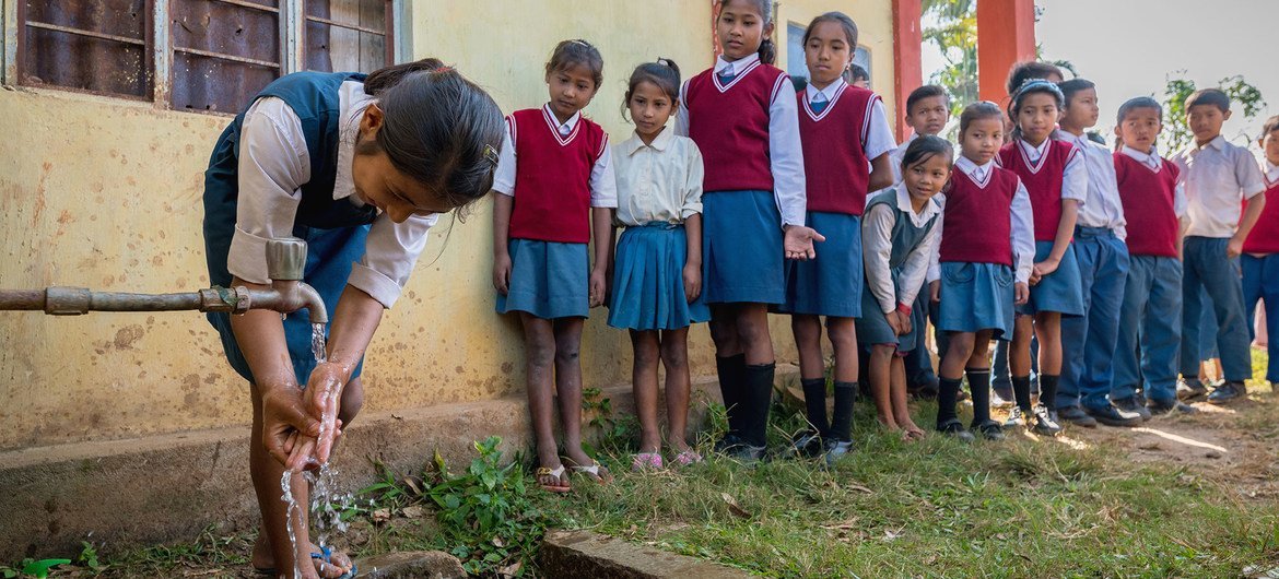 स्कूल की वर्दी पहने एक छात्रा बाहर लगे नल पर हाथ धो रही है, जबकि वर्दी पहने अन्य बच्चे कतार में खड़े होकर उसे देख रहे हैं।