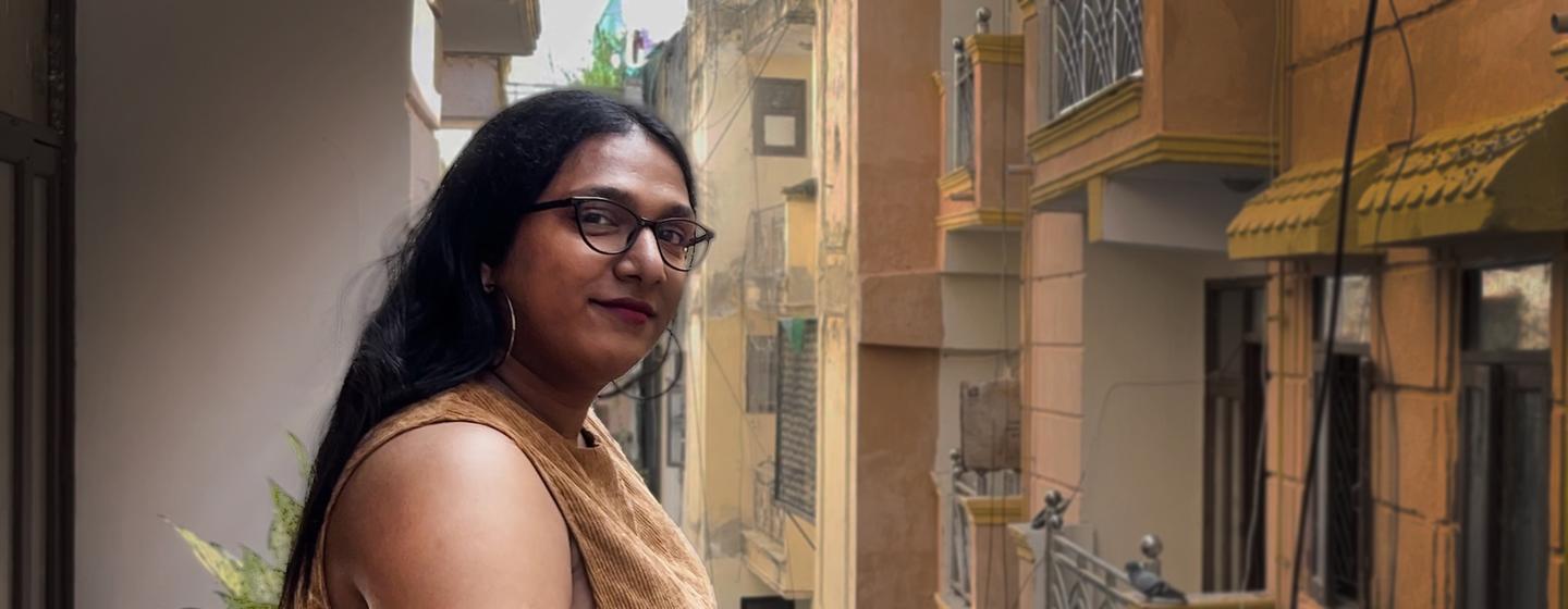 भारत में एक संकरी शहरी गली के सामने एक बालकनी पर चश्मा और भूरे रंग की पोशाक पहने एक ट्रांसजेंडर महिला खड़ी है।