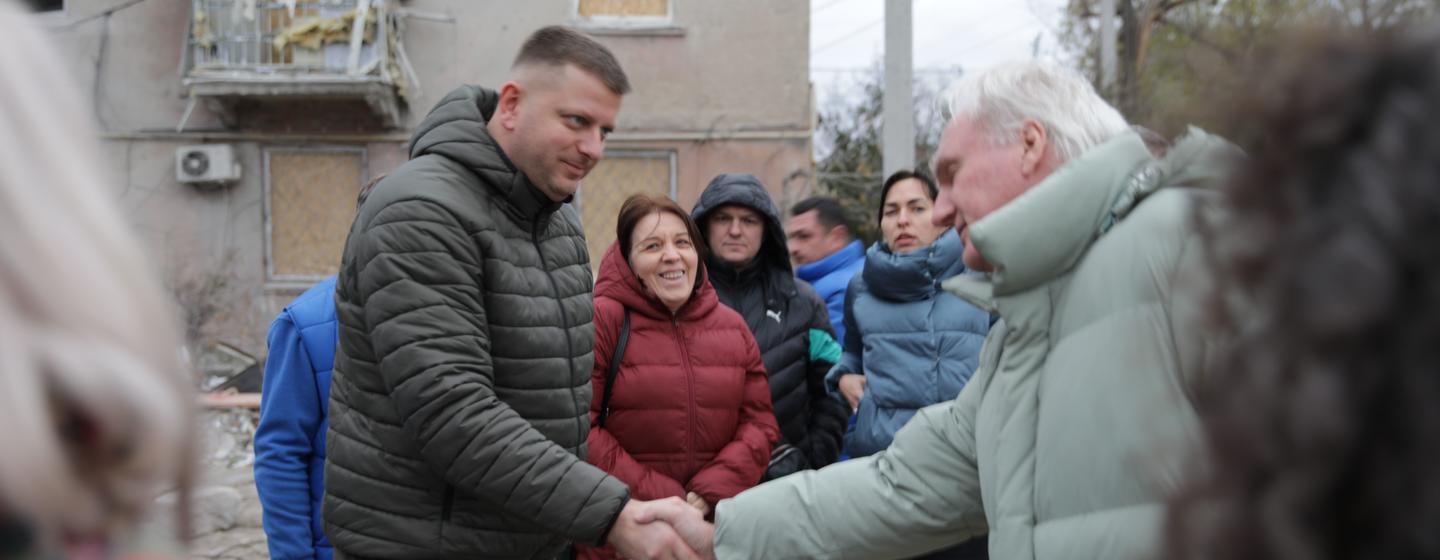 Un homme vêtu d'une doudoune sombre serre la main d'un homme plus âgé portant un manteau vert clair, entouré d'un petit groupe de personnes devant un bâtiment endommagé en Ukraine.