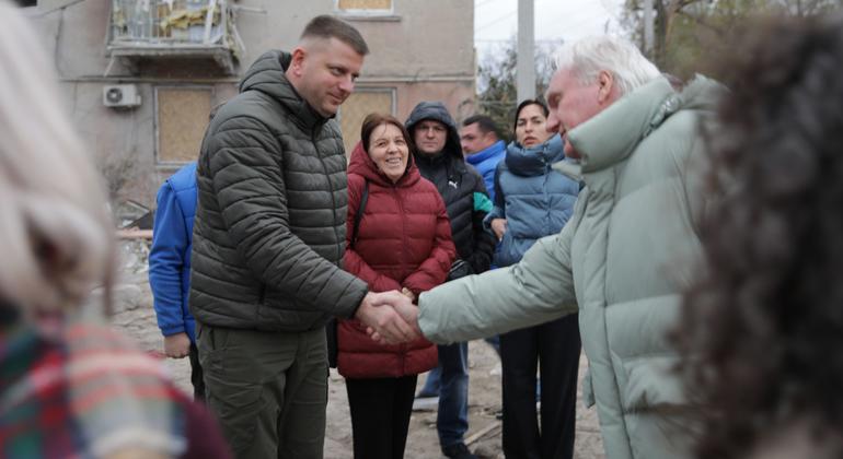Мужчина в темной пуховой куртке пожимает руку пожилому мужчине в светло-зеленом пальто в окружении небольшой группы людей перед поврежденным зданием на Украине.