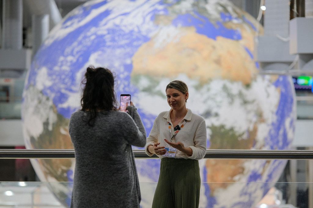Женщина выступает на открытии мероприятия Together4Transparency в рамках COP30 в Белеме, Бразилия, а другой человек фотографируется на фоне большого глобуса Земли.