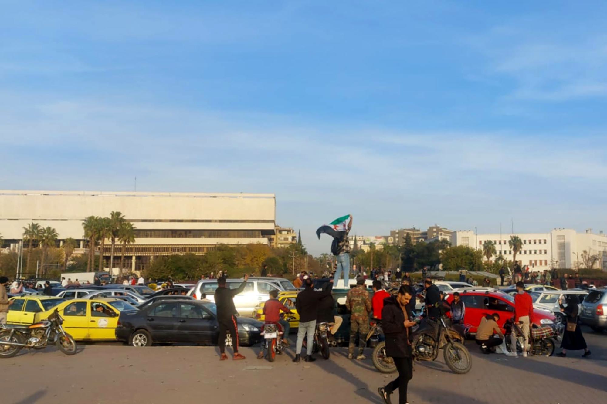يحتفل الناس في ساحة الأمويين بدمشق، سوريا، وسط السيارات والدراجات النارية التي تحوم حولهم والعلم السوري يرفرف.