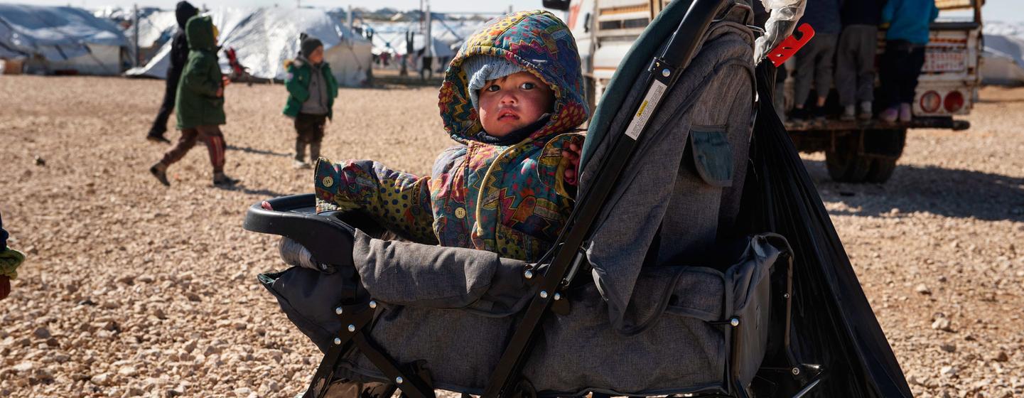 Uma criança pequena está sentada em um carrinho de bebê no campo de refugiados de Al-Hol, no nordeste da Síria, com tendas e um caminhão ao fundo.