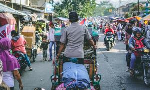 A man pedals a traditional Becak rickshaw down a busy street in Bandung City, Indonesia.