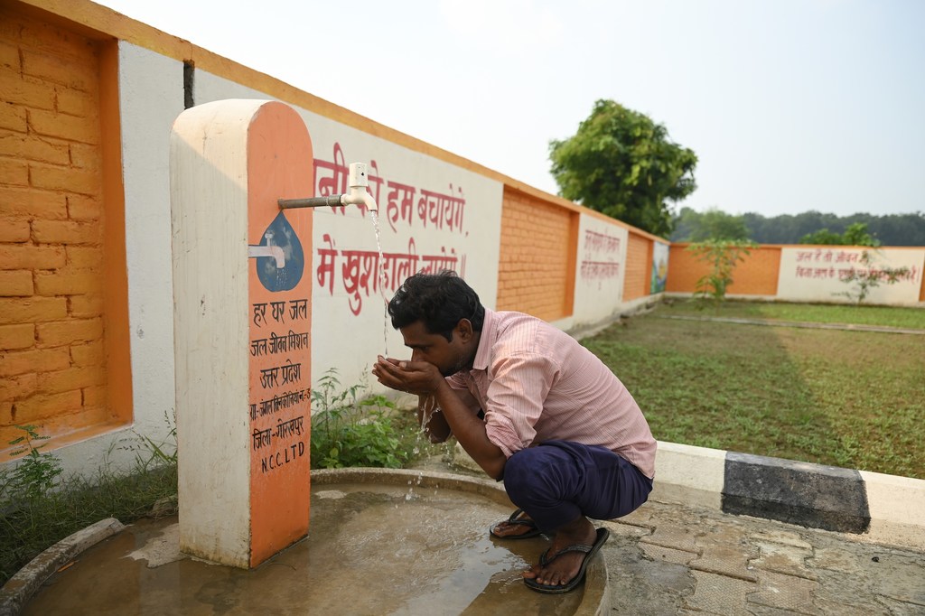 एक व्यक्ति सामुदायिक जल केंद्र पर सार्वजनिक नल से पानी पी रहा है, जिसकी दीवार पर स्वच्छ जल और स्वच्छता के लिए UNICEF इंडिया दस्तक कार्यक्रम को बढ़ावा देने वाला लेख लिखा हुआ है।