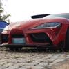 Um Toyota Supra vermelho está estacionado em uma rua de paralelepípedos, com outro carro visível ao fundo.