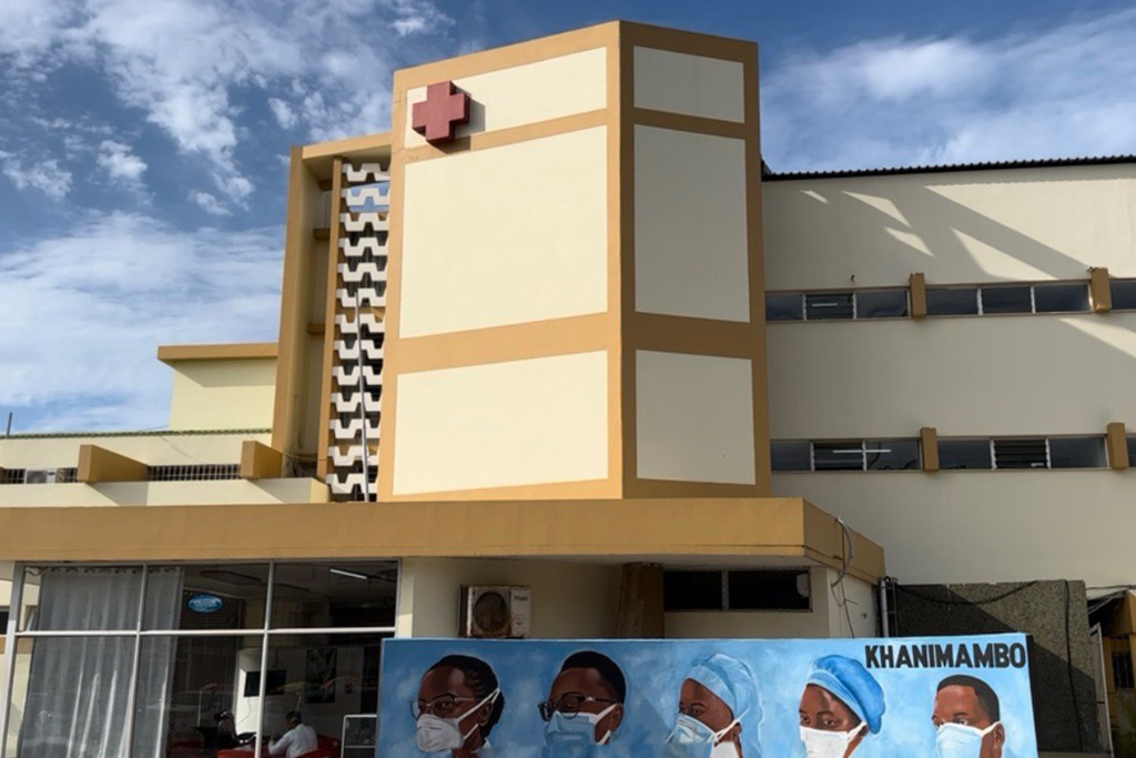 Vista exterior de um edifício hospitalar em Moçambique com o símbolo da cruz vermelha e uma faixa com cinco profissionais de saúde.
