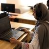 Афганская женщина в платке и наушниках работает за ноутбуком за столом.