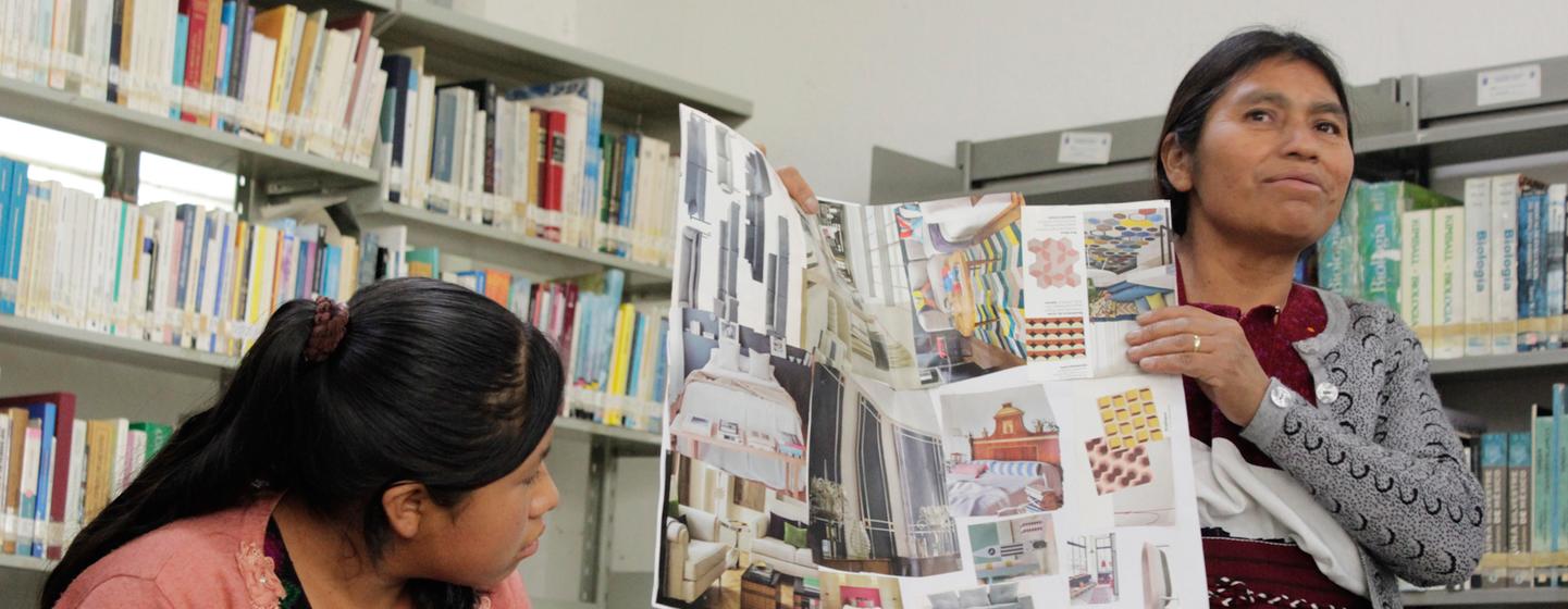 Una mujer muestra un portafolio de trabajos de diseño de interiores a otra mujer en una biblioteca o taller.