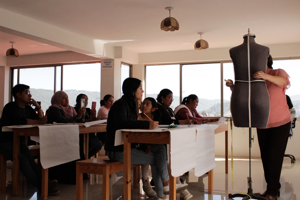 Un grupo de personas se encuentra en un aula, donde un instructor les muestra cómo utilizar un maniquí para la confección de prendas.