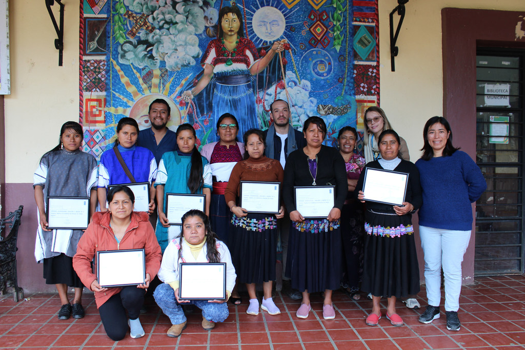 Un grupo de mujeres artesanas de Chiapas, México, sosteniendo certificados frente a un colorido mural.