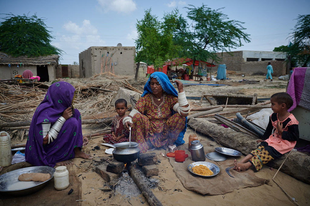 Dos mujeres y dos niños se sientan frente a sus casas dañadas en una aldea de Pakistán, preparando una comida sobre una hoguera. La mujer del centro remueve la olla mientras los demás la rodean. Al fondo se aprecian escombros y estructuras dañadas.