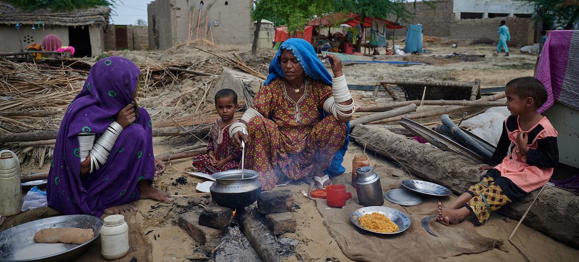 پاکستان کے ایک گاؤں میں دو خواتین اور دو بچے اپنے تباہ شدہ گھروں کے باہر بیٹھ کر کھلی آگ پر کھانا بنا رہے ہیں۔ بیچ میں موجود عورت ایک برتن کو ہلاتی ہے جبکہ دیگر اس کے ارد گرد بیٹھتے ہیں۔ پس منظر میں ملبہ اور تباہ شدہ ڈھانچے نظر آ رہے ہیں۔