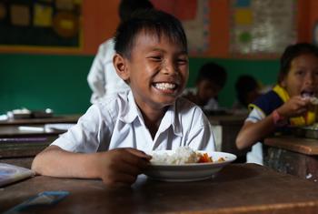 Um jovem aluno cambojano sorri alegremente em uma sala de aula enquanto come um prato de arroz fortificado.