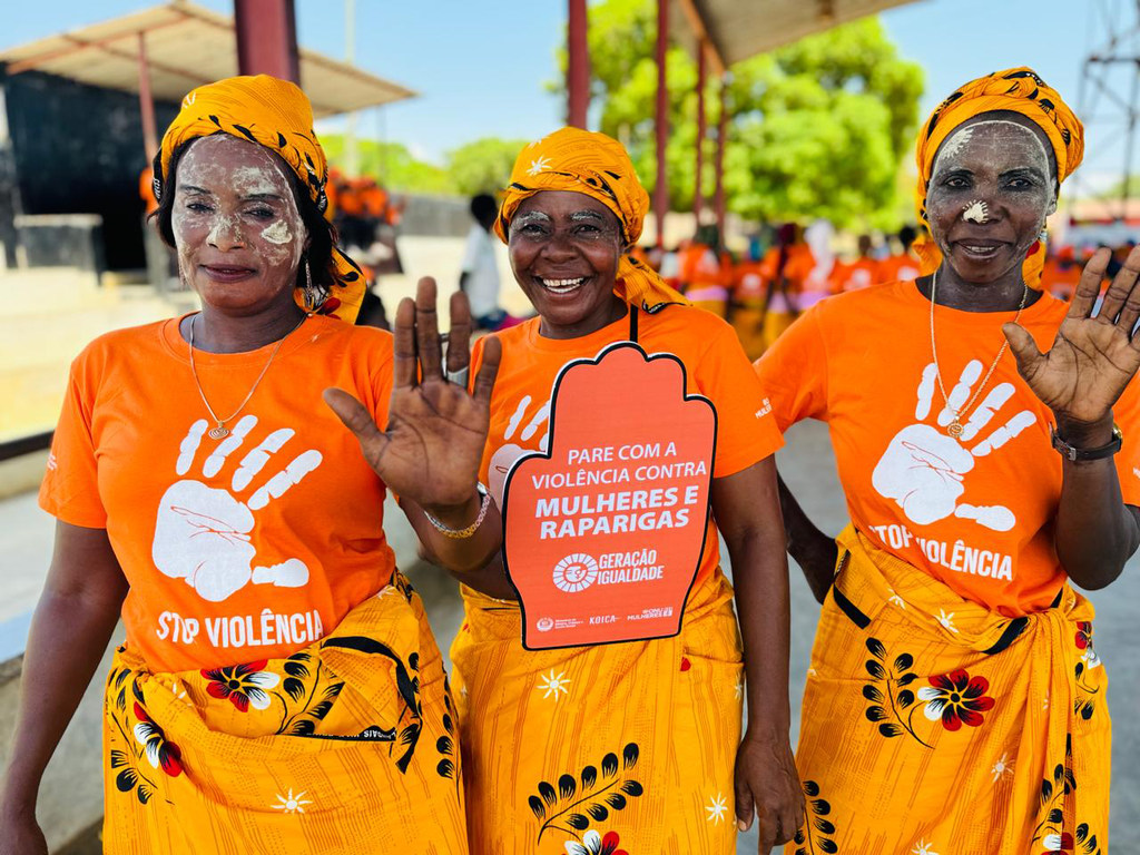 Três mulheres vestidas de laranja e com lenços na cabeça participam de um protesto em Moçambique, segurando uma placa com os dizeres "Parem a violência contra mulheres e meninas".