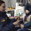 Женщина в синей куртке с нашивкой в ​​виде украинского флага кормит маленького ребенка в синей шапке и пальто в помещении, которое, по всей видимости, является временным убежищем.
