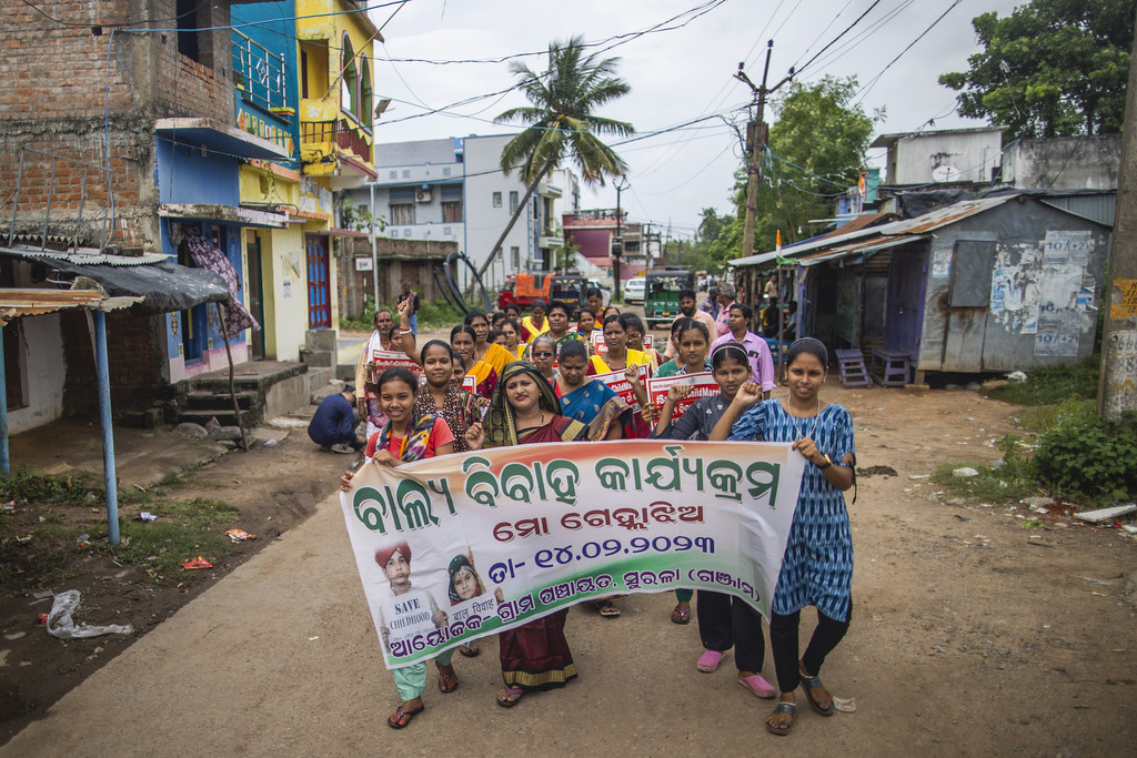 बाल विवाह को रोकने के लिए आयोजित एक सामुदायिक रैली में भारतीय महिलाओं का एक समूह ग्रामीण सड़क पर एक बड़ा बैनर पकड़े हुए भाग ले रहा है।