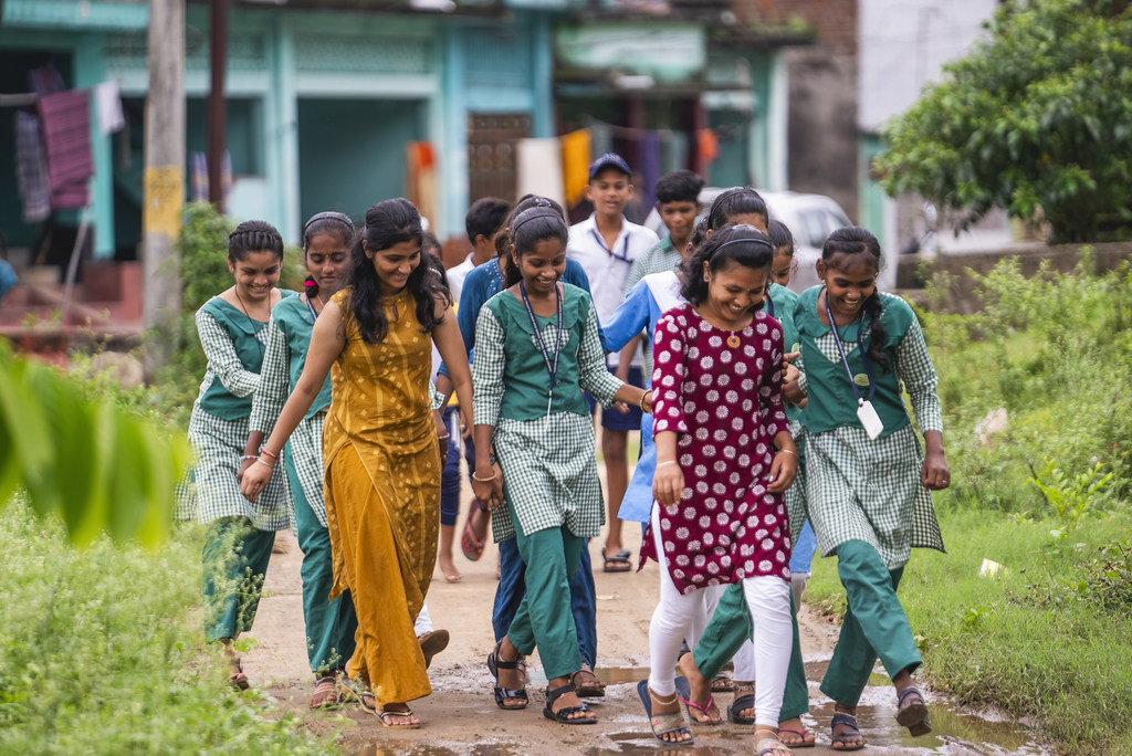 भारत के ओडिशा में स्कूल की वर्दी पहने युवा लड़कियों का एक समूह एक वयस्क महिला के साथ ग्रामीण रास्ते पर चल रहा है।