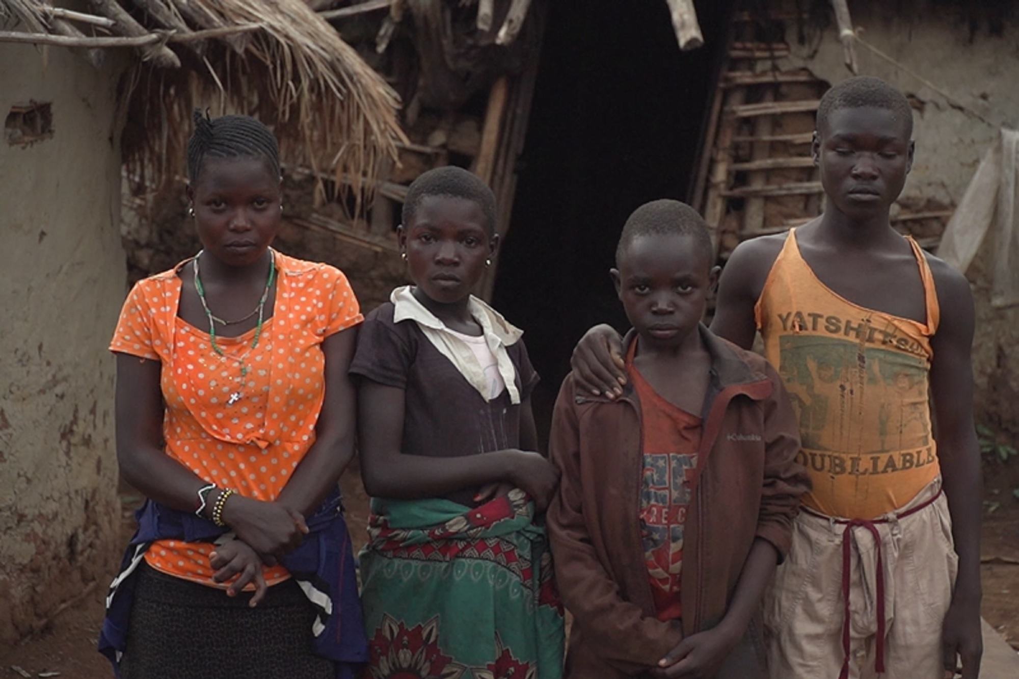 Watoto wanne wadogo wa Kiafrika wamesimama kwa safu mbele ya nyumba rahisi yenye paa la nyasi na kuta za matope katika mazingira ya vijijini.