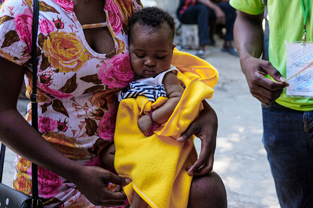 Una mujer con un vestido estampado de flores sostiene a un bebé desnutrido envuelto en una manta amarilla en un centro de nutrición de UNICEF en Puerto Príncipe, Haití.