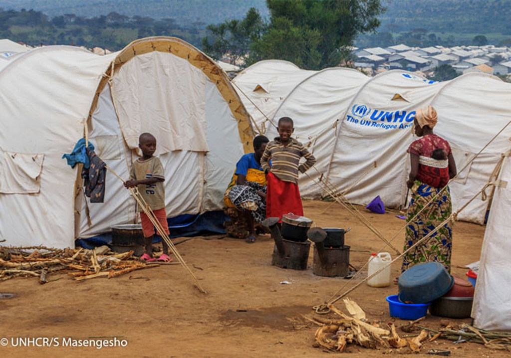 تعيش عائلة في خيمة تابعة UNHCR في مخيم للاجئين في رواندا. يتجمع الأطفال والبالغون في الخارج، مع وضع أواني الطبخ على الموقد والحطب على الأرض.