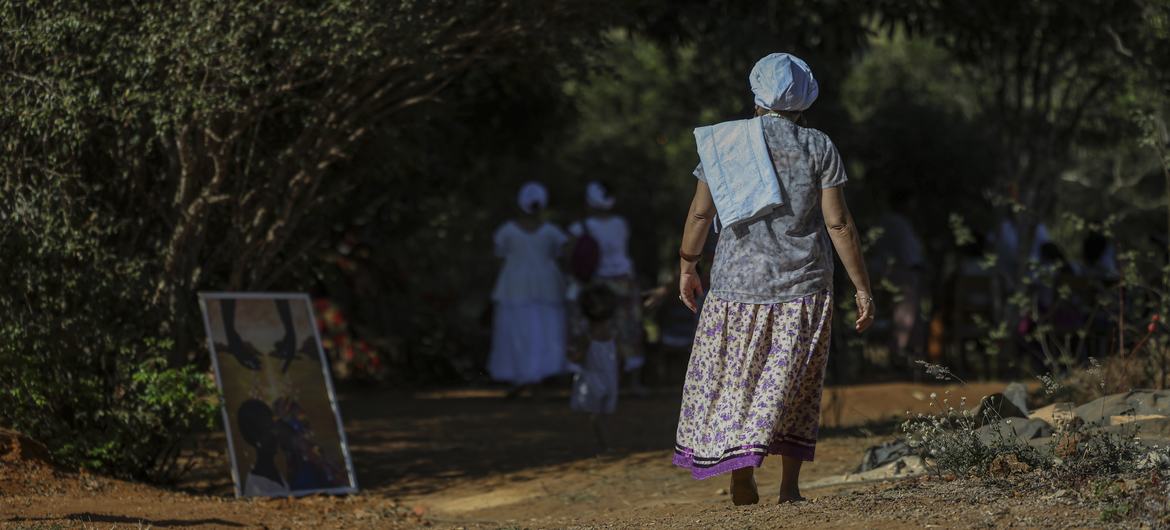 Uma mulher vestida com trajes tradicionais caminha por uma trilha de terra durante um festival, com outras pessoas e árvores ao fundo.