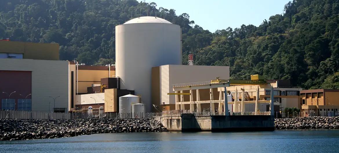 A usina nuclear Angra 1, no Rio de Janeiro, Brasil, com seu edifício de contenção e estruturas industriais circundantes, tendo como pano de fundo uma colina arborizada e ao lado de um corpo d'água.