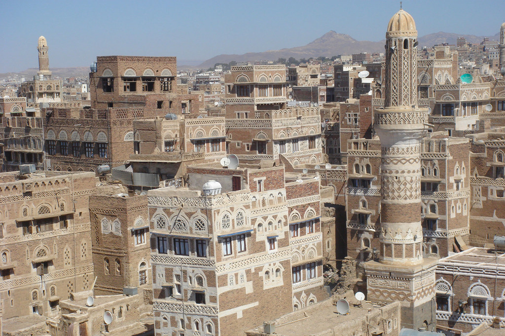 Vista panorámica de la Ciudad Vieja de Sana'a, Yemen, que muestra edificios tradicionales de adobe con intrincadas tallas y minaretes prominentes con las montañas como telón de fondo.