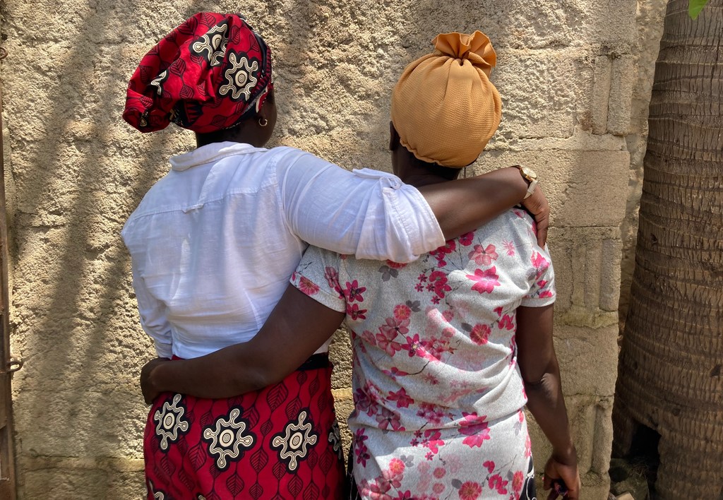 Duas mulheres estão de pé, com os braços em volta dos ombros uma da outra, vistas por trás, em Moçambique.
