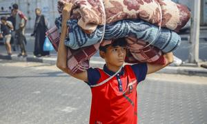 A 14-year-old boy carries mattresses and blankets on his head while his family follows him to an emergency shelter in the Gaza Strip.