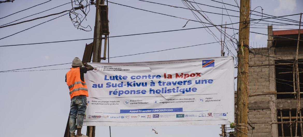 Mfanyakazi aliyevaa fulana ya rangi ya chungwa akiweka bango kubwa kuhusu kampeni ya uhamasishaji ya Mpox katika jamii huko Kivu Kusini, DR Congo.
