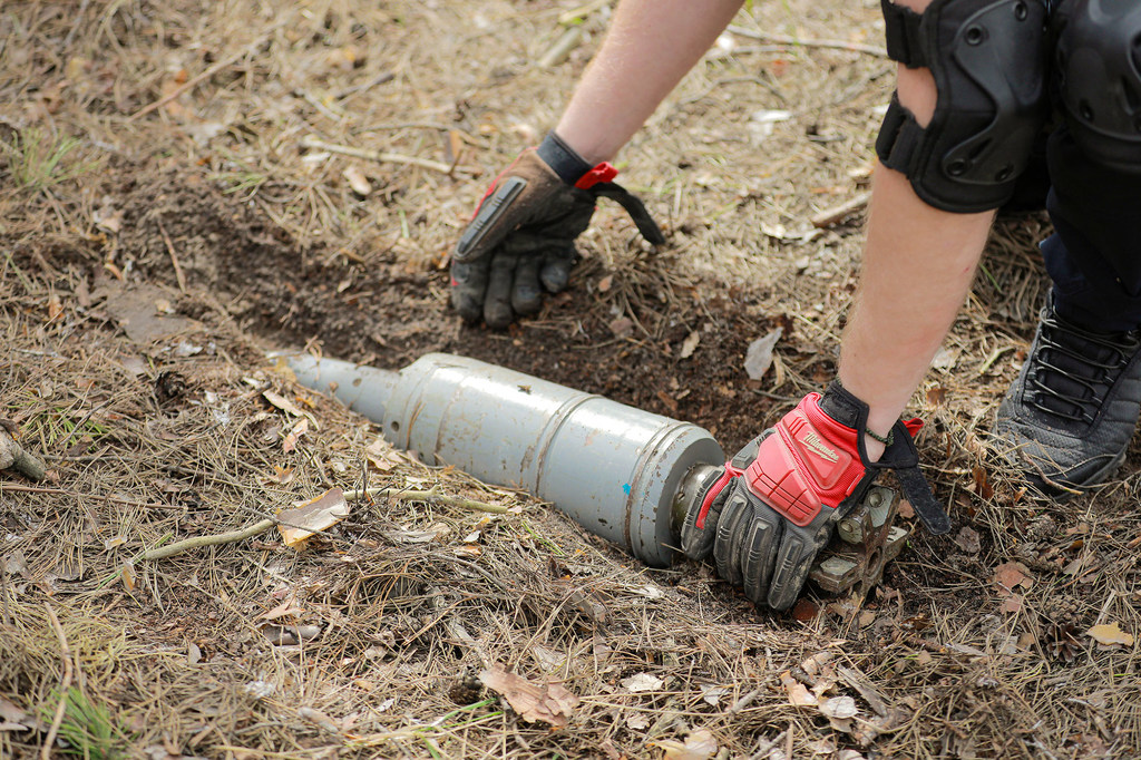 A deminer's hands in protective gloves and knee pads carefully handle an unexploded landmine in a forest.