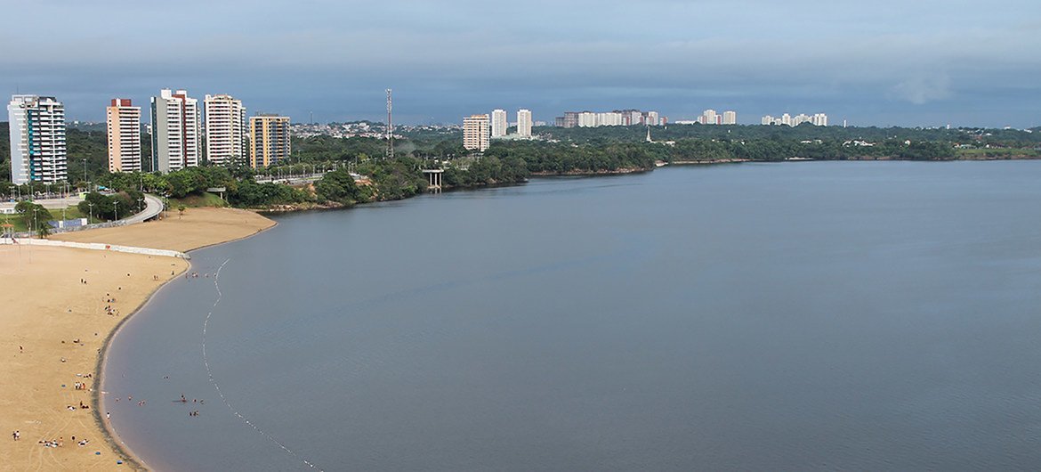 Vista panorâmica de uma praia de areia às margens de um grande rio em Manaus, Brasil, com edifícios altos ao fundo.