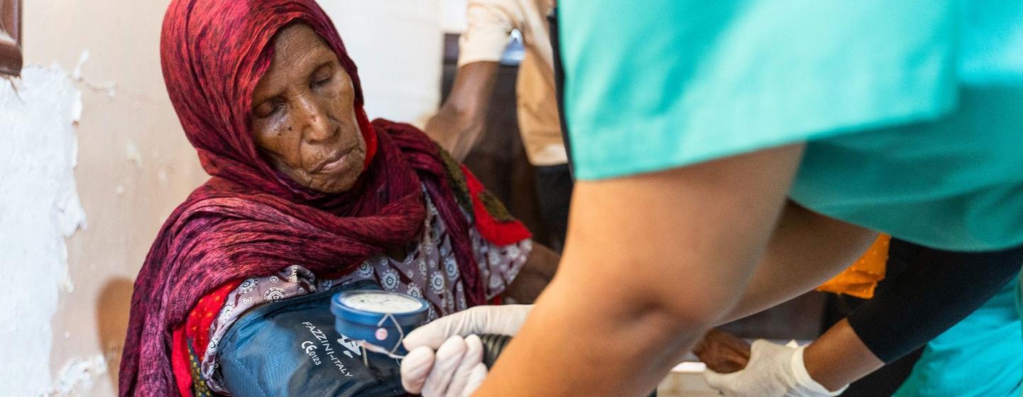 Dans un cadre clinique, un professionnel de santé en tenue verte mesure la tension artérielle d'une femme âgée portant un foulard rouge.
