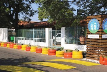 La entrada de una escuela en Paraguay cuenta con un paso de peatones marcado con pintura amarilla para carreteras y una hilera de neumáticos pintados de colores que sirven como macetas. Se puede ver un letrero de madera con el logotipo "mi camino escolar seguro".