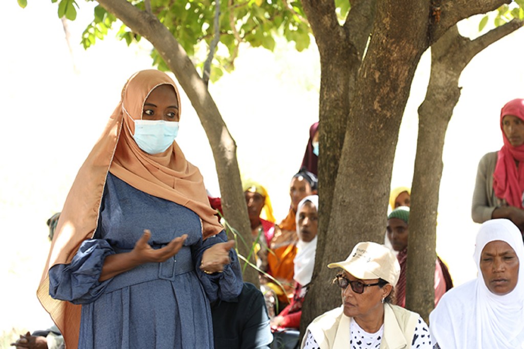 Mwanamke aliyevaa hijab na barakoa anazungumza na kundi la watu nje chini ya mti.