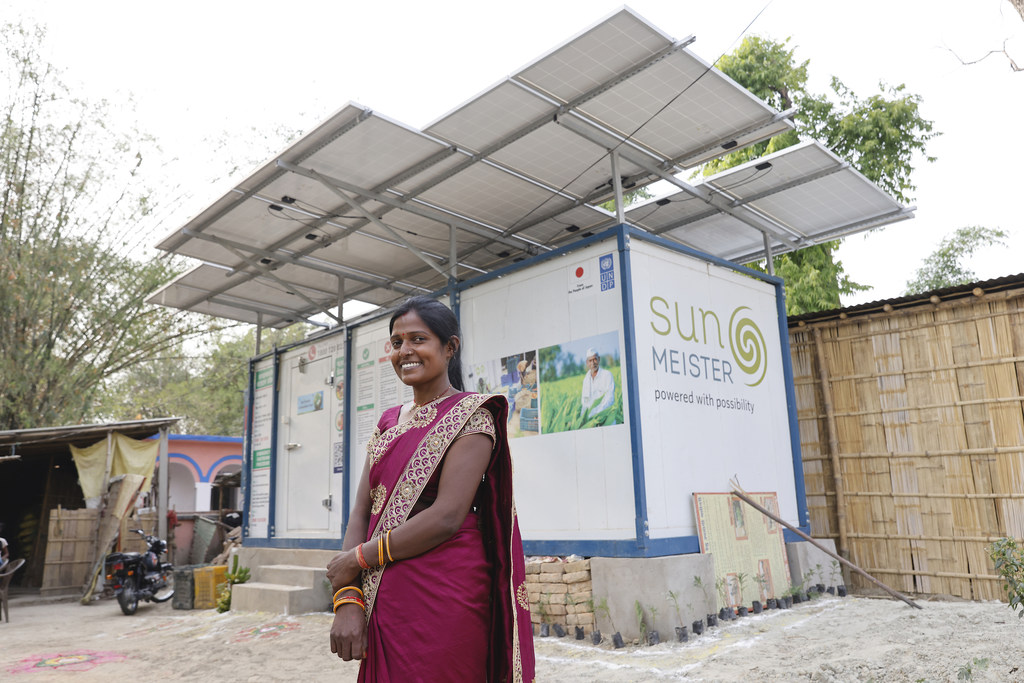 एक मुस्कुराती हुई भारतीय महिला UNDP इंडिया की परियोजना 'सन मीस्टर' के लोगो वाली सौर ऊर्जा से चलने वाली कोल्ड स्टोरेज यूनिट के सामने खड़ी है।