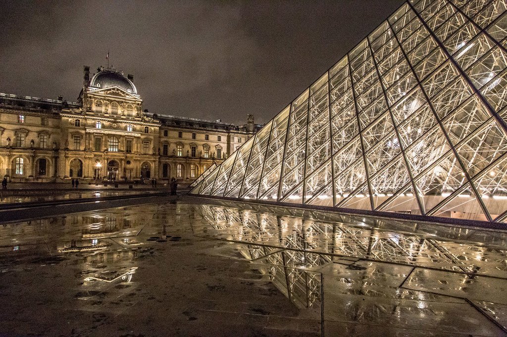 El Museo del Louvre iluminado y su pirámide de cristal por la noche en París, con sus reflejos visibles en el pavimento mojado del patio.