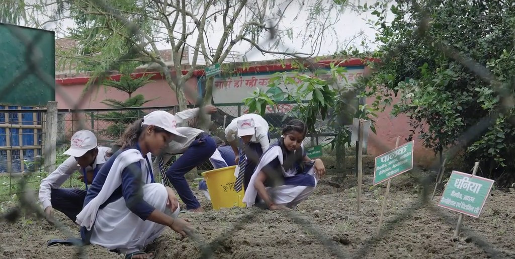 भारतीय स्कूली छात्राएं वर्दी पहने एक बगीचे में झुकी हुई हैं, प्लास्टिक कचरे को छांट रही हैं और गीले कचरे से खाद बना रही हैं।