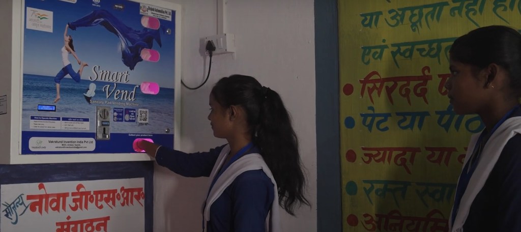 स्कूल यूनिफॉर्म पहने एक लड़की सैनिटरी पैड बेचने वाली मशीन चला रही है।